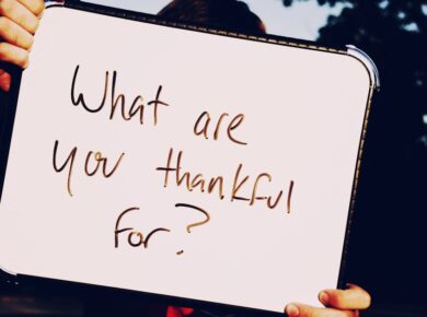 A person holds a whiteboard asking 'What are you thankful for?' against a dark outdoor background.