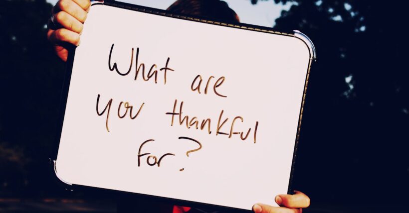 A person holds a whiteboard asking 'What are you thankful for?' against a dark outdoor background.