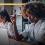 Two women working at laptops in a modern office space, focused on their tasks.