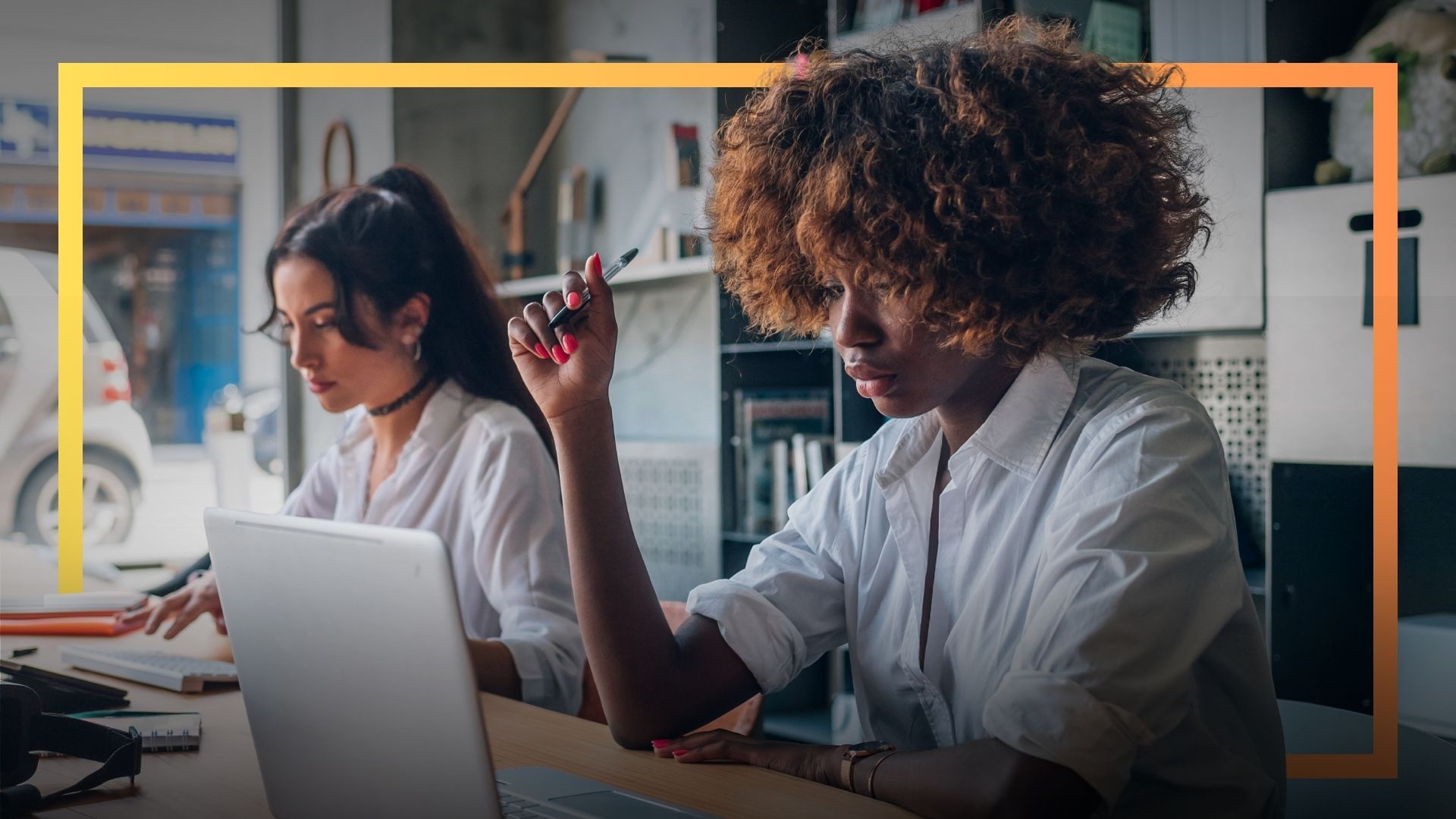 Two women working at laptops in a modern office space, focused on their tasks.